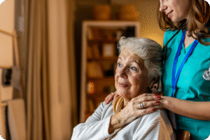 A healthcare professional comforts a senior woman in a cozy, well-lit room with bookshelves in the background.