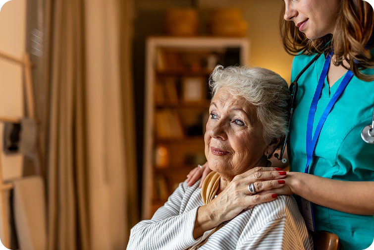 A healthcare professional comforts a senior woman in a cozy, well-lit room with bookshelves in the background.