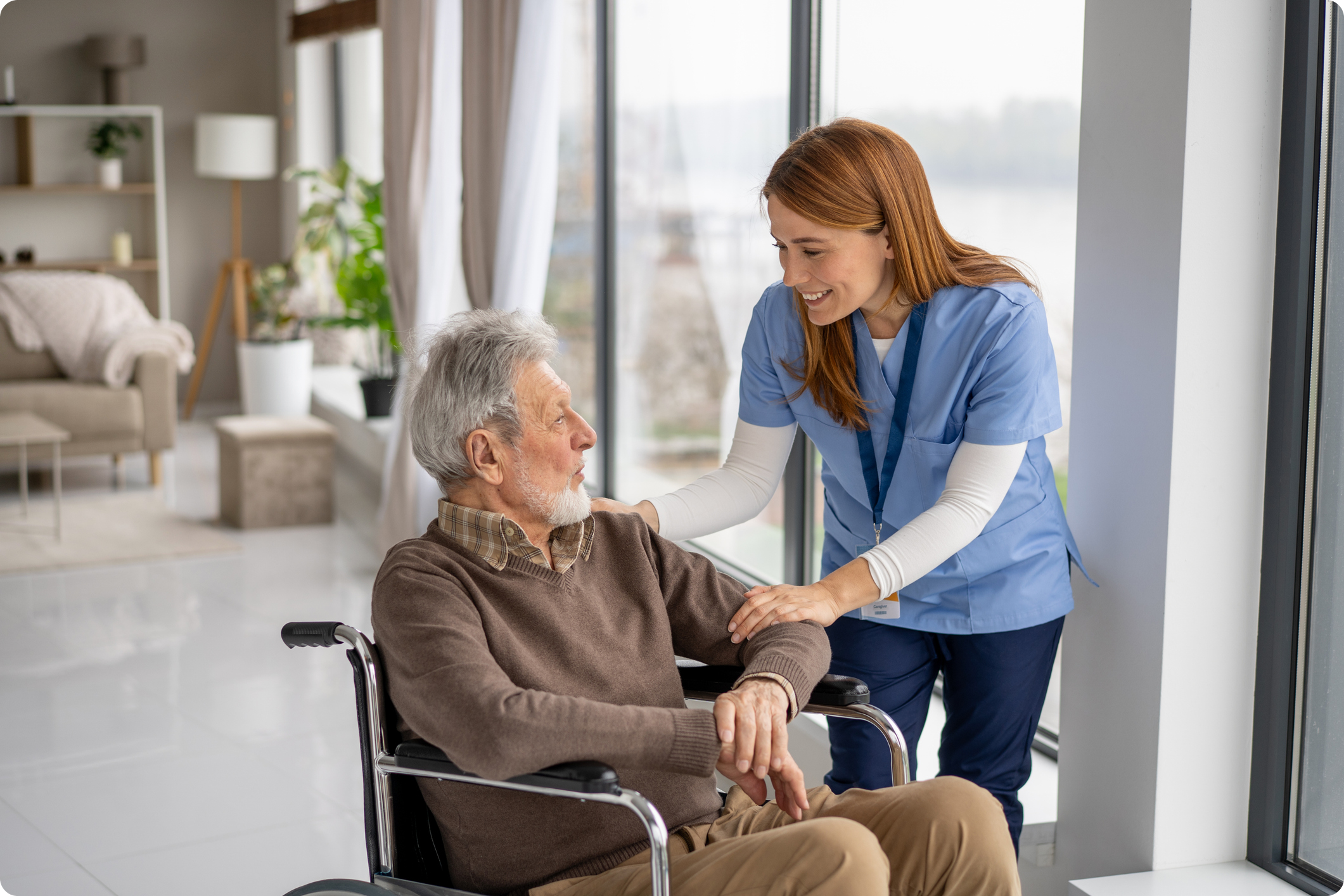 A nurse in scrubs converses with an elderly man in a wheelchair, set in a bright, modern healthcare environment.