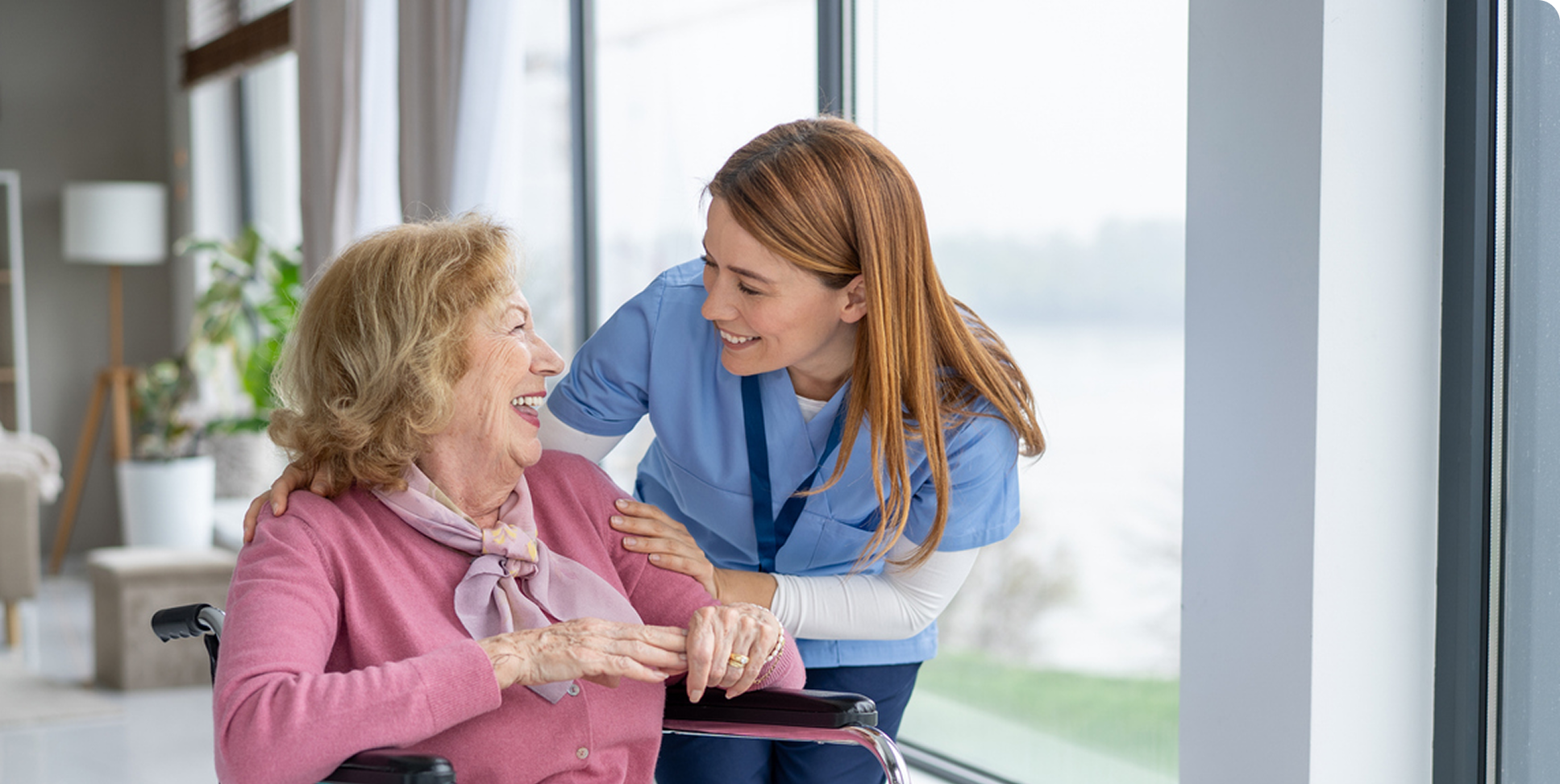 A caregiver smiles while assisting an elderly woman in a wheelchair in a bright, modern healthcare environment.