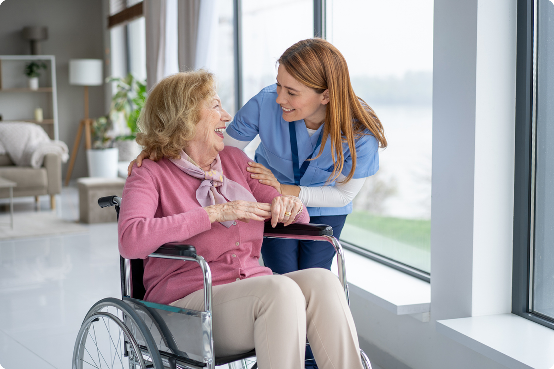 A caregiver smiles while assisting an elderly woman in a wheelchair in a bright, modern healthcare environment.