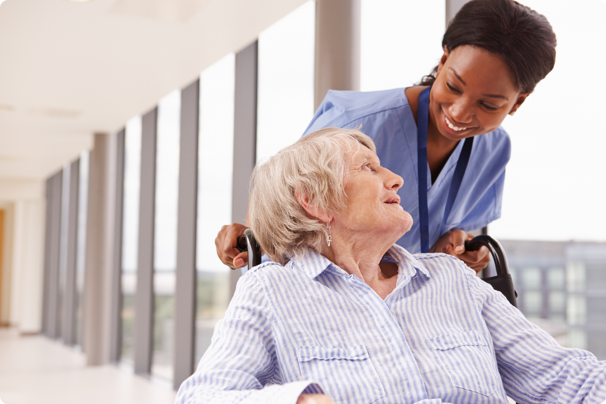 A smiling nurse assists an elderly woman in a wheelchair in a bright, modern healthcare facility corridor.