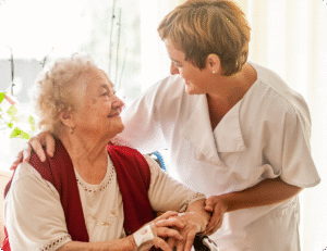 A caregiver interacts warmly with an elderly woman in a bright room filled with plants and soft natural light.