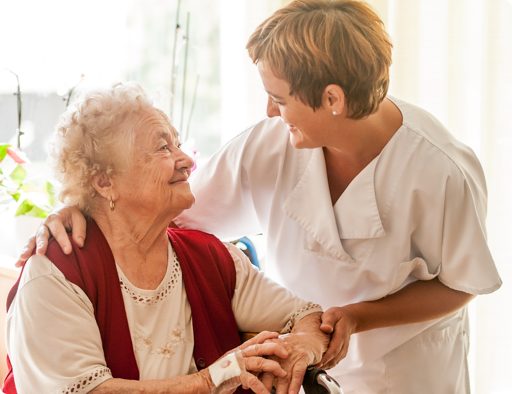 A caregiver interacts warmly with an elderly woman in a bright room filled with plants and soft natural light.