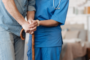 A healthcare professional assists an older adult using a wooden cane in a comfortable interior setting.
