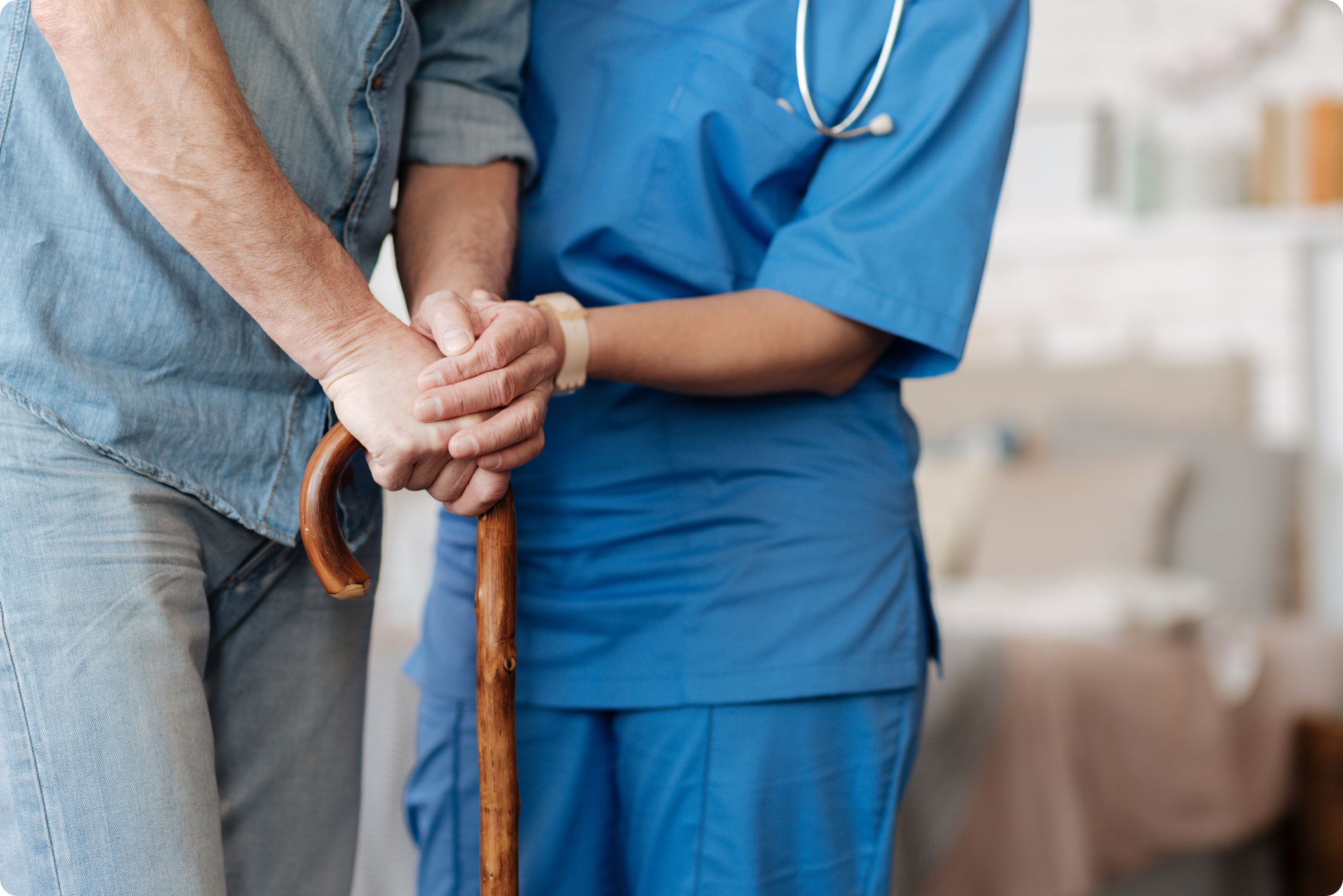 A healthcare professional assists an older adult using a wooden cane in a comfortable interior setting.
