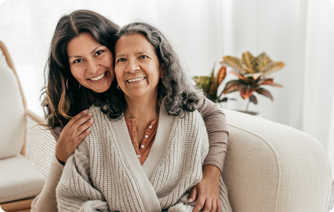A smiling young woman embraces an elderly woman in a cozy, well-lit indoor setting with potted plants in the background.