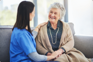 A caregiver and an elderly woman share a warm moment on a couch in a bright, inviting living space.
