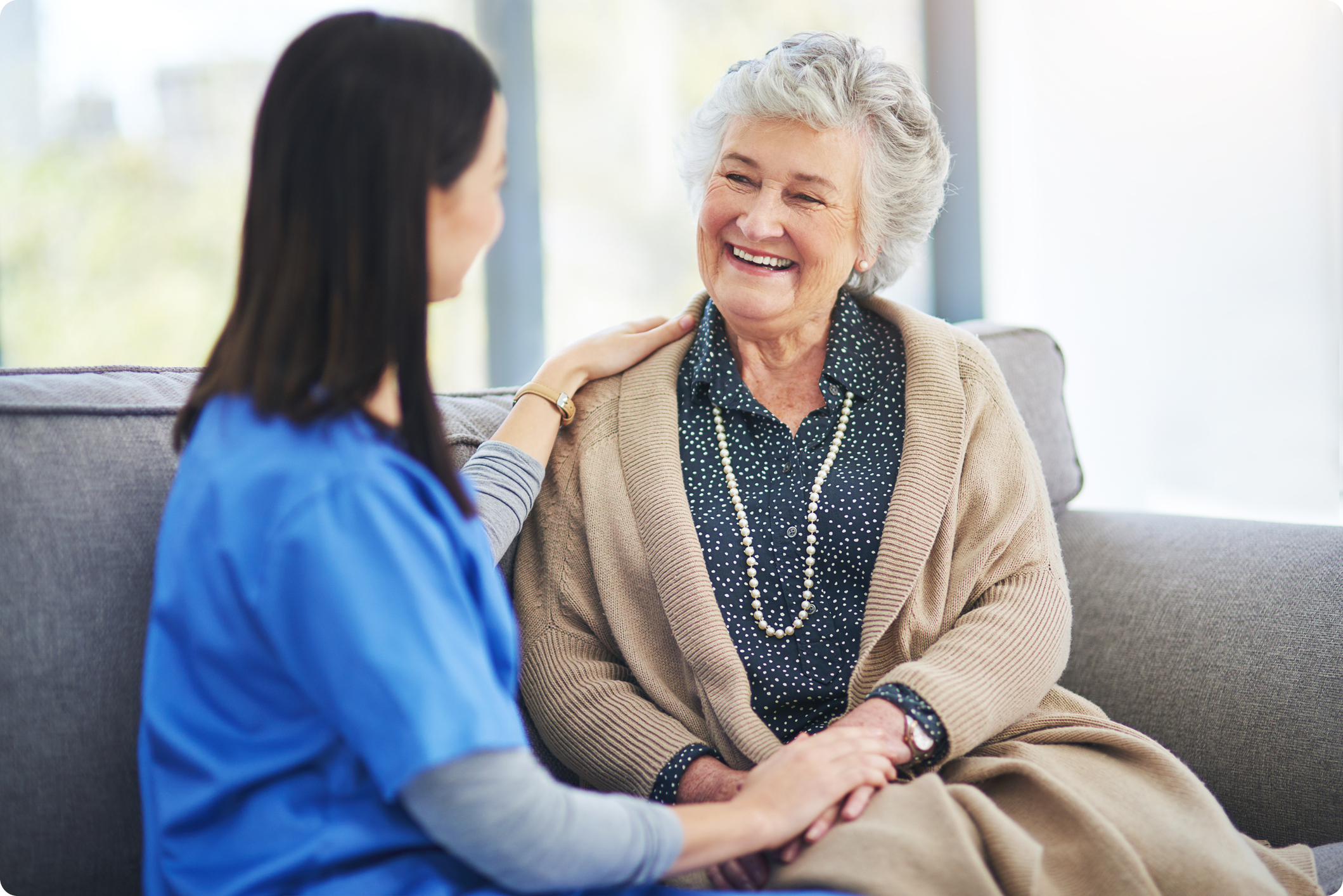 A caregiver and an elderly woman share a warm moment on a couch in a bright, inviting living space.