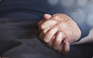 Two hands, one aged and the other younger, clasped together gently against a soft fabric background.