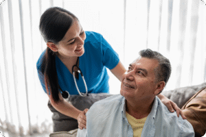 Nurse in blue scrubs engages with an older man wearing glasses, both smiling in a well-lit, comfortable indoor setting.