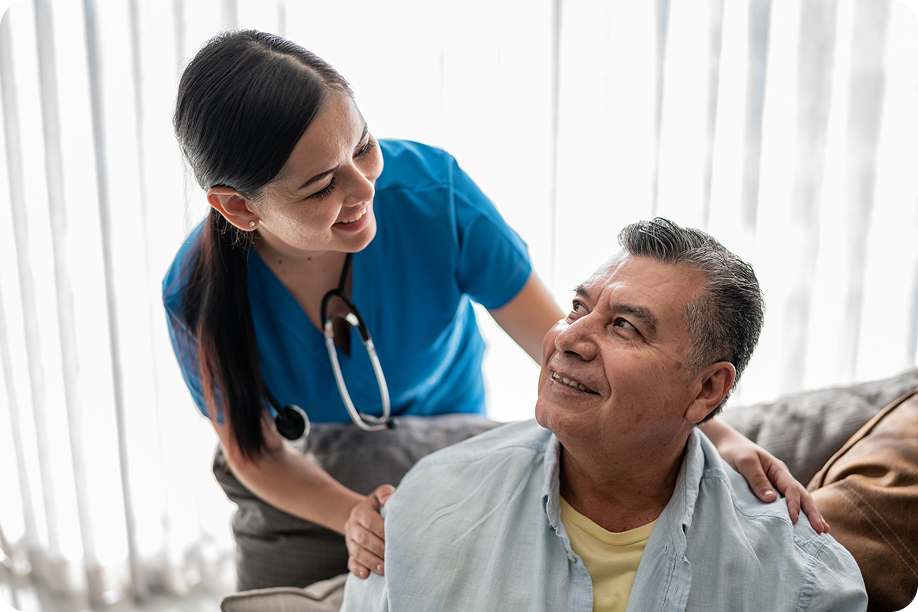 Nurse in blue scrubs engages with an older man wearing glasses, both smiling in a well-lit, comfortable indoor setting.