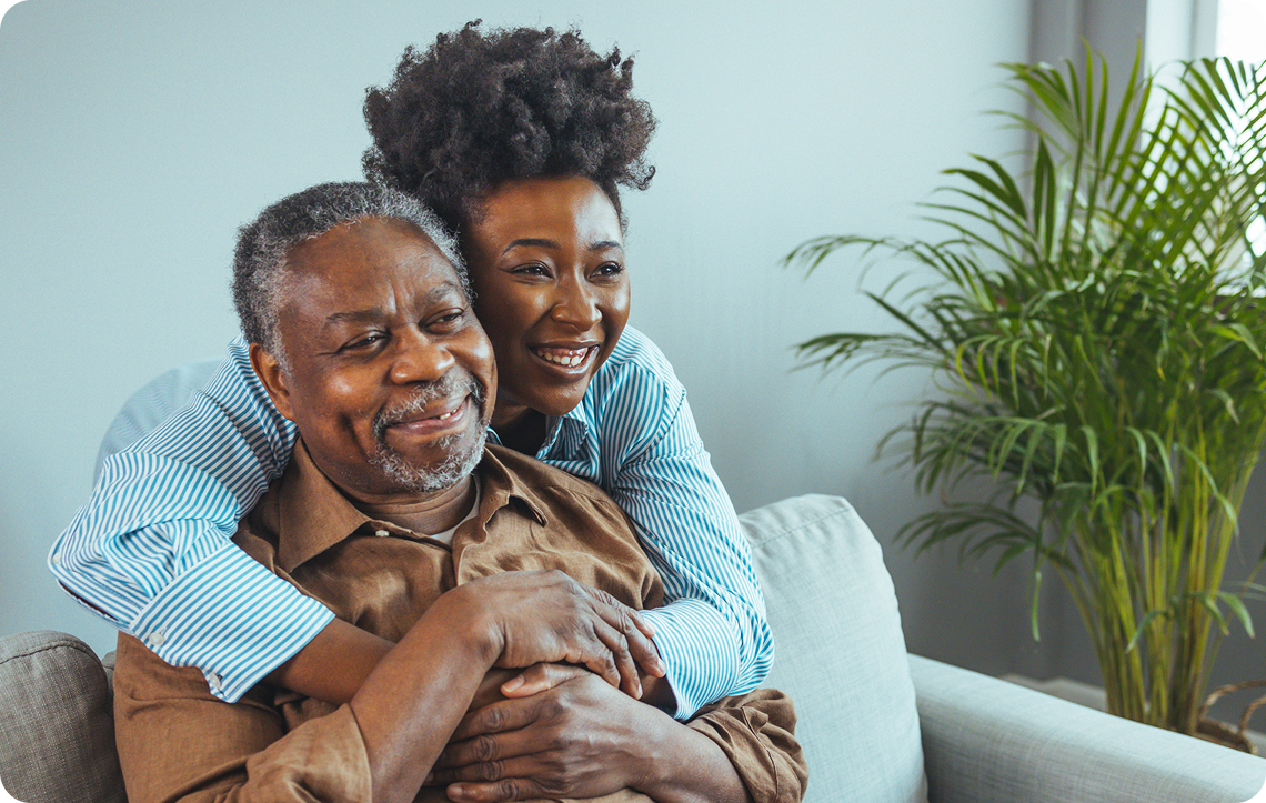 A smiling woman embraces an older man while sitting on a couch, with indoor plants in the background.