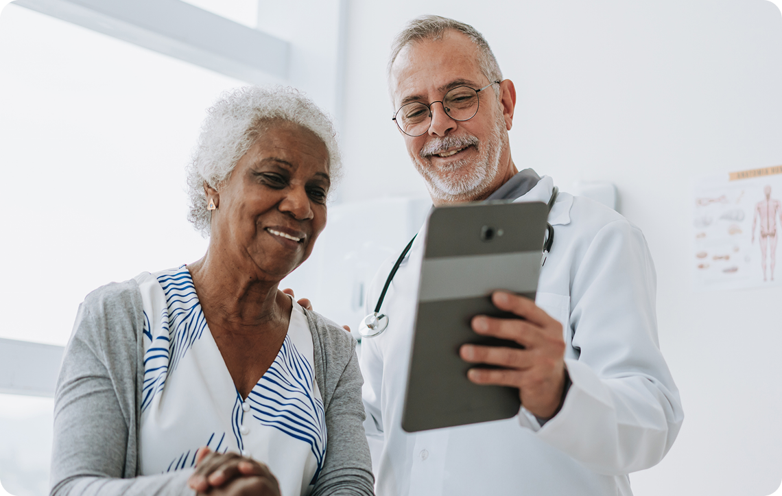 A friendly, middle-aged doctor in a white coat shows a smiling senior woman a tablet.