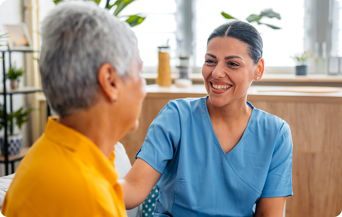 A happy female caregiver in blue scrubs smiles at a senior woman with gray hair.