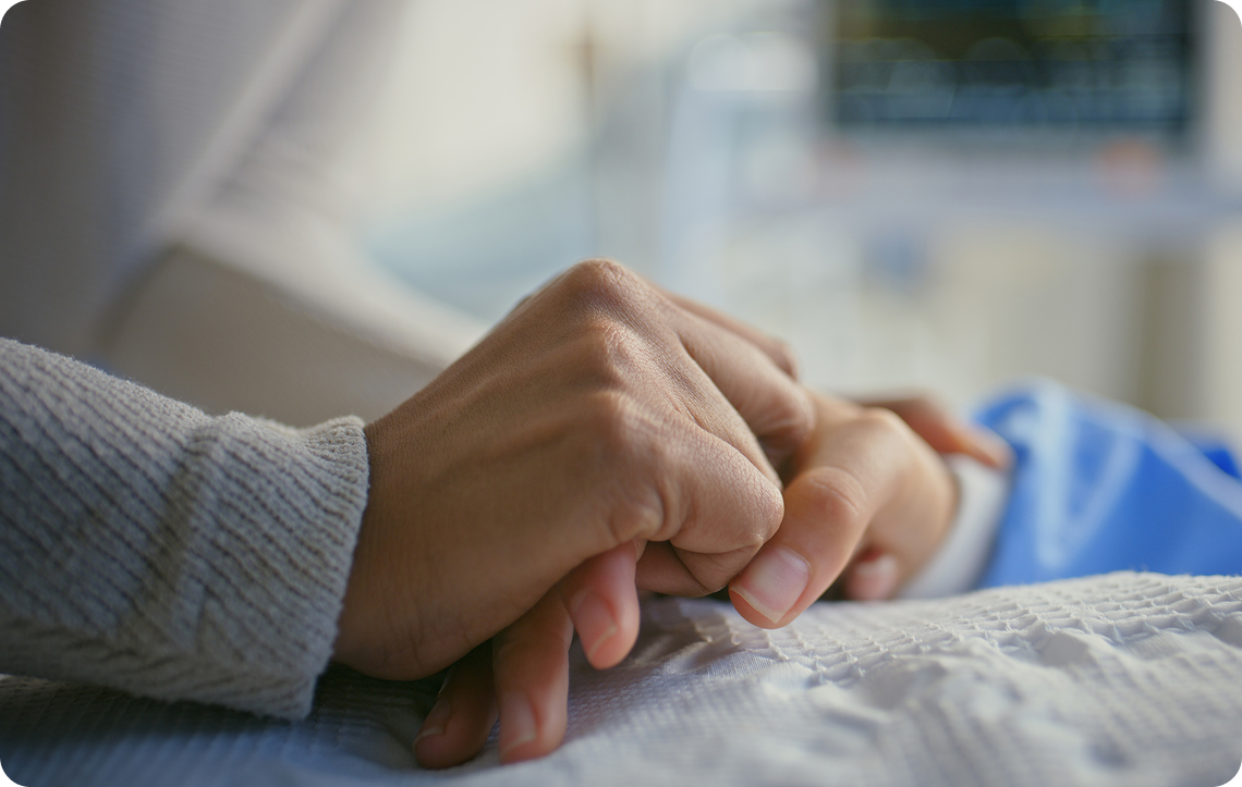 Two hands, one aged and the other younger, clasped together gently against a soft fabric background.