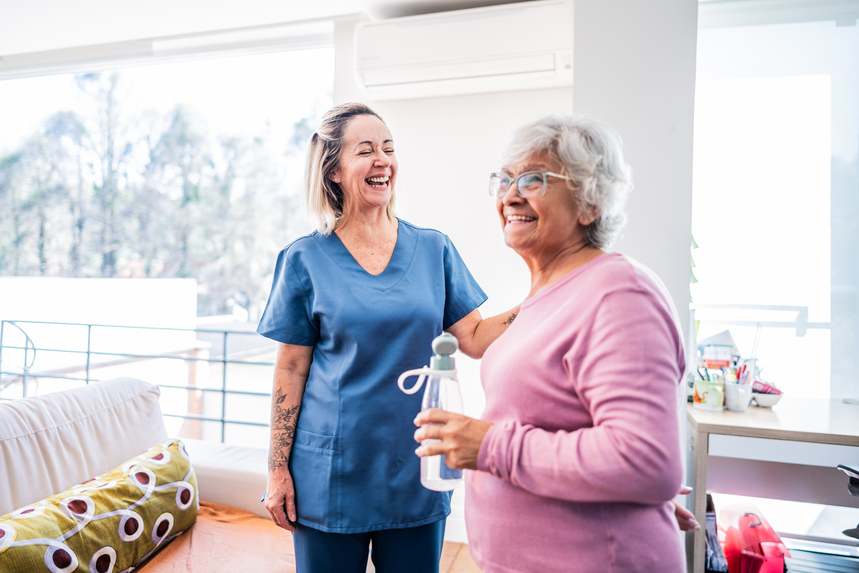 Two women, a nurse in scrubs and an elderly woman, share a joyful moment in a bright, modern room with natural light.